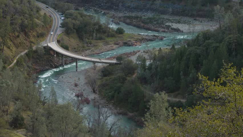 A curved bridge crosses a turquoise river, connecting two shores surrounded by dense forest. The top-down view highlights the winding road path and the natural harmony of the landscape