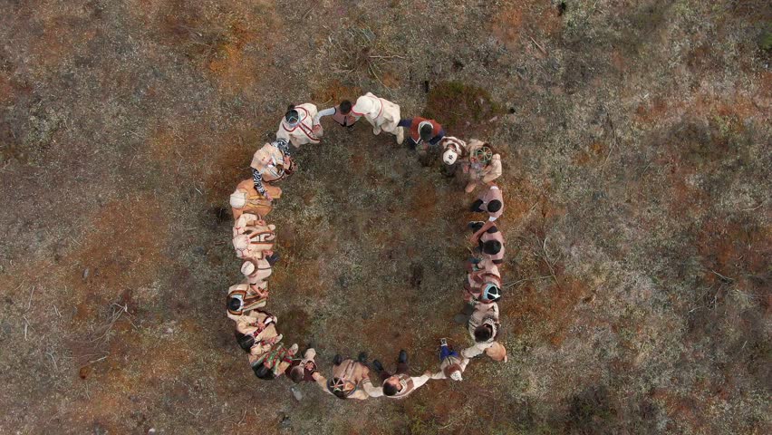 Captivating aerial footage of a nomadic tribe in vibrant national costumes performing a circular dance amidst the vast, desolate summer tundra beyond the Arctic Circle. The drone captures the harmony 