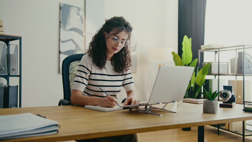 Curly-haired woman wearing glasses sitting at office desk and writing in notebook. Holding pen while taking notes, concentrating on task. Appearing engaged and very thoughtful about task.