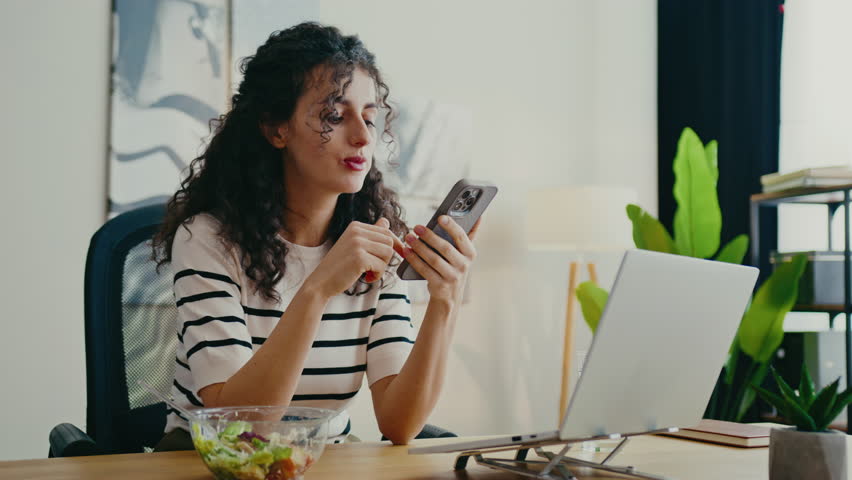 Caucasian girl checking smartphone while eating salad. Holding fork, smiling slightly while reading screen. Checking notifications or reading message from friend. Relaxed atmosphere at workspace.