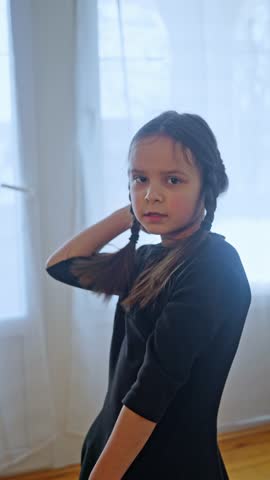 The child dances and smiles. A young girl dressed in a beautiful black dress with lovely braids is found in a bright room. Slow motion