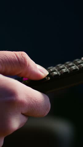 Vertical video Close up of man in dimly lit apartment using remote control to browse though movies on streaming service. Remote control used by person in living room flipping through channels on TV