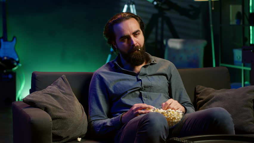 Fatigued man watching shows on TV late at night and munching on popcorn, paying close attention to screen. Tired sedentary man sitting in pajamas all day consuming mindless entertainment, camera A