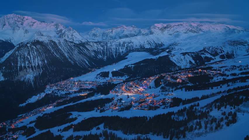 Experience the breathtaking and stunning aerial view over Courchevel, France, at night, where the sparkling snowy mountains meet a charming village that is beautifully illuminated by colorful lights