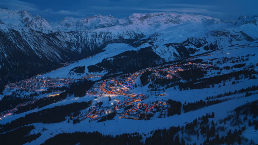 Aerial view of Courchevel, France, at night,where glowing village streets and ski slopes shine against majestic snow-covered mountains. Gentle snowfall enhances enchanting winter wonderland atmosphere