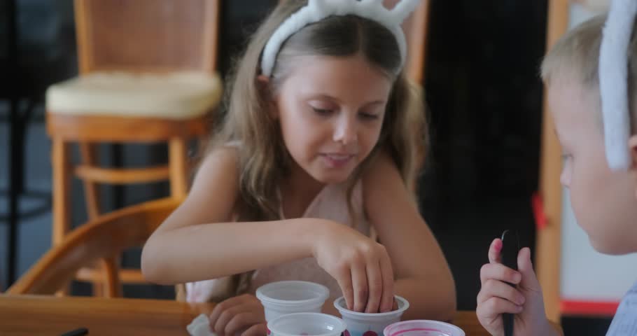 Children at bunny ear headbands sit at the kitchen and paint eggs in different colors for the holiday of Holy Easter. They have fun and dip eggs in different cups with paint. Preparation for holiday.