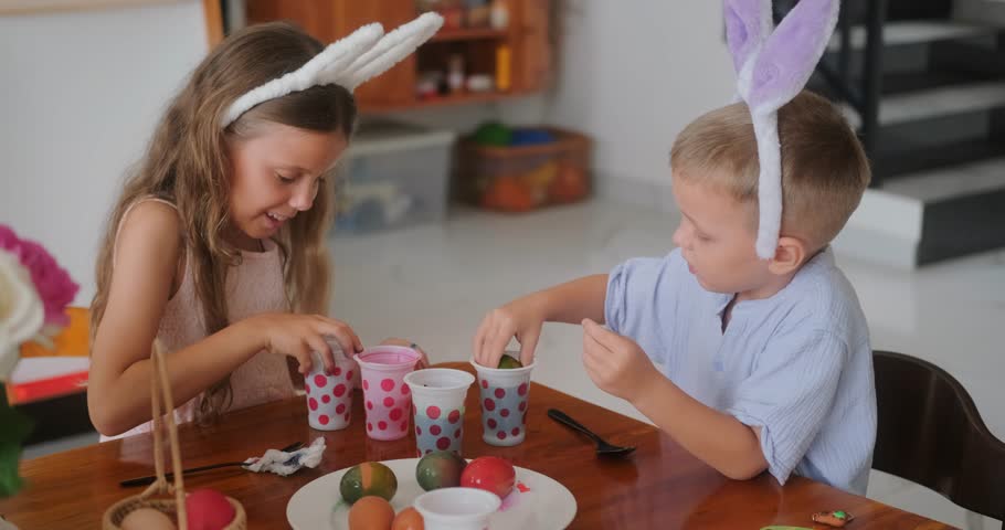 Children at bunny ear headbands sit at the kitchen and paint eggs in different colors for the holiday of Holy Easter. They have fun and dip eggs in different cups with paint. Preparation for holiday.