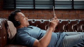 Locked medium shot, Side view of Asian man lying on sofa and use tablet and headphone to listening music Or surf on internet - Powered by Shutterstock - Get 15% off with code: PIKWIZARD15