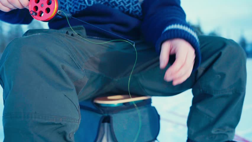 A Man is Spinning the Reel of an Ice Fishing Rod While Sitting on a Stool in a Snowy Environment - Close Up