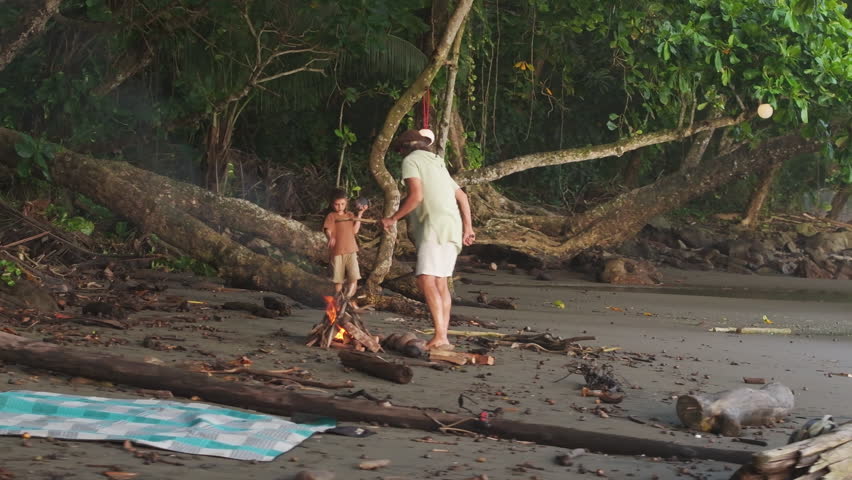 A serene aerial view shows a father and son lighting a warm fire on a tranquil Costa Rican beach, surrounded by lush greenery and soft sands, creating unforgettable memories in nature