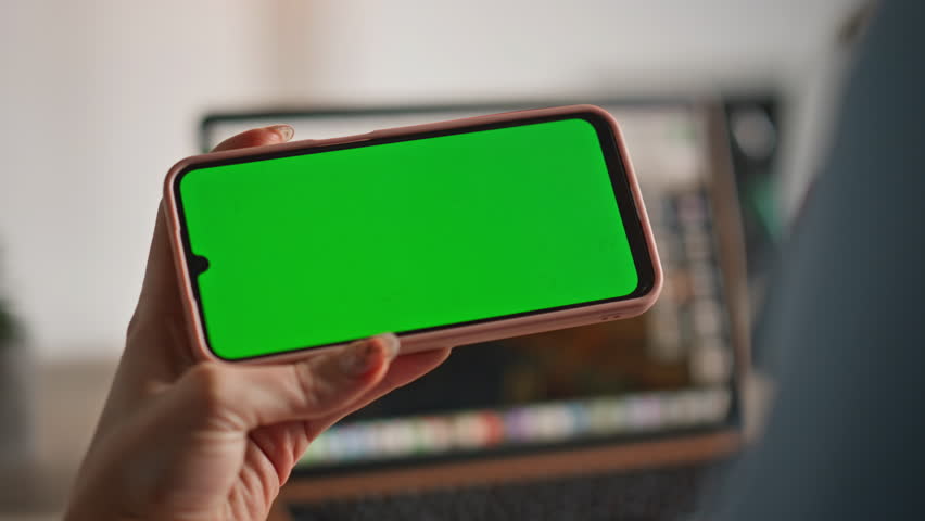 Woman watching video at mockup mobile phone at home office closeup. Unknown female freelancer browsing social media on green screen smartphone. Businesswoman hands hold horizontal chroma key cellphone