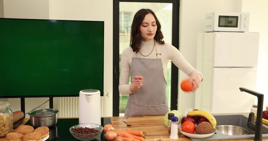 Pretty young woman in apron plays with fresh aromatic oranges in kitchen. Brunette female cooks tasty organic salad with exotic fruits in sunny room