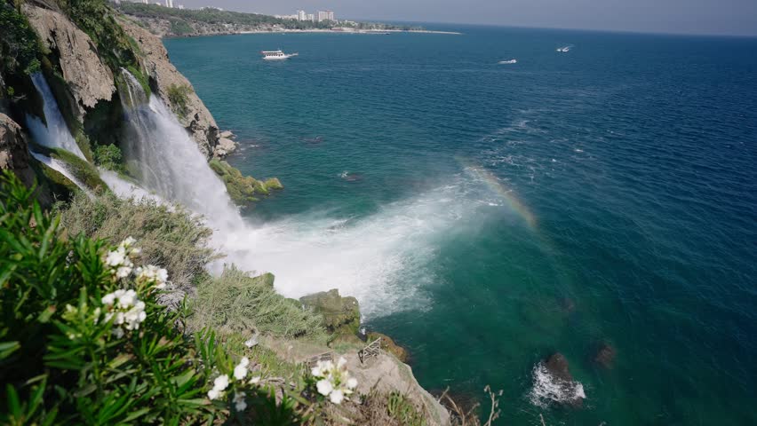 Powerful Lower Duden Waterfall plunging dramatically into Mediterranean Sea, generating sparkling white foam and vibrant rainbow against scenic coastal landscape near Antalya, Turkey