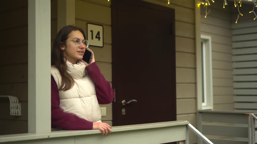A woman enjoys a phone call on her porch, decorated with beautiful holiday lights