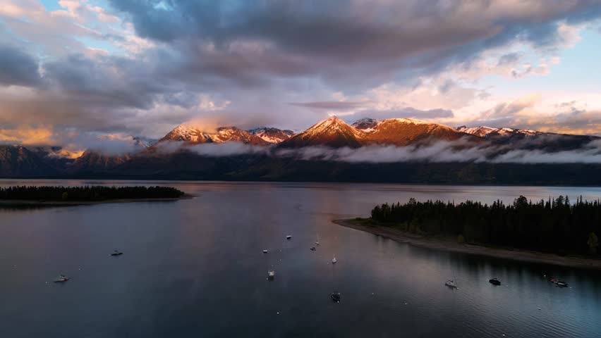 Aerial view of serene sunrise over a picturesque lake with a sailboat, snowy mountains, and lush forest islands, Grand Teton, Wyoming, United States.
