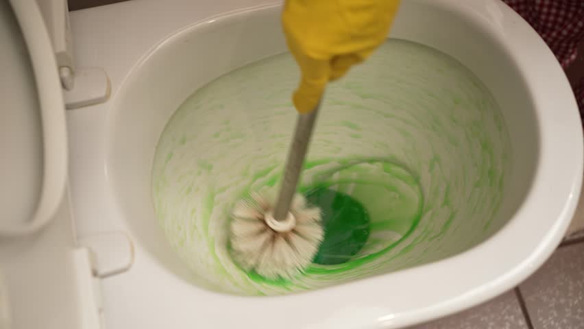 Green disinfectant streaming into toilet bowl, gloved hand scrubbing ceramic surface with toilet brush, ensuring thorough sanitization and pristine bathroom hygiene