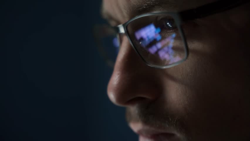 Close up of focused software engineer or data scientist wearing glasses, analyzing code on his screen. Programming script reflects on his eyeglasses as he works late.