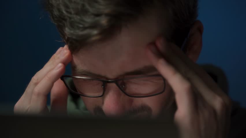A tired coder wearing eyeglasses working late at night feels a headache and rubs his eyes from fatigue. Overwork concept