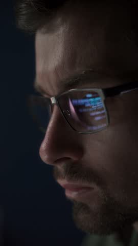 Close up of data scientist face is closely examines data. The glow of code lines reflecting in software engineer glasses and eyes. Vertical video