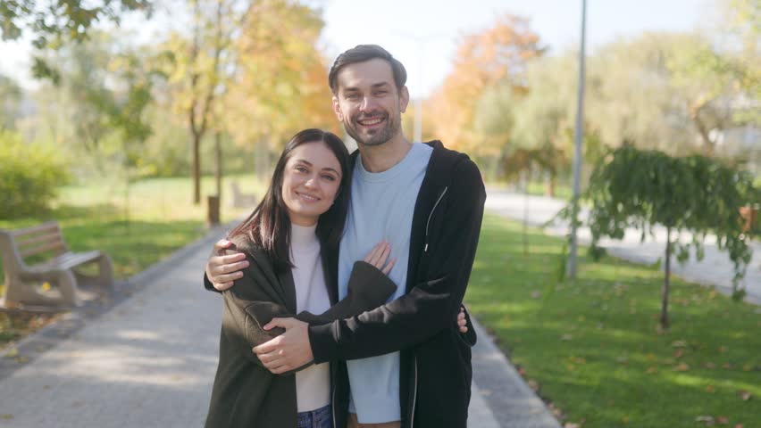 A young couple dressed in casual autumn attire embraces lovingly in a scenic park. Surrounded by colorful leaves, they smile warmly at the camera, embodying the spirit of autumn romance.