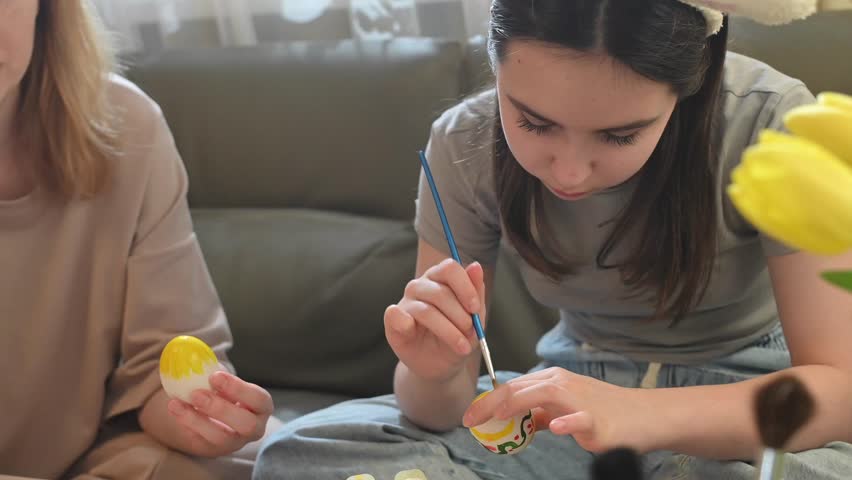 Mother and daughter painting Easter eggs together at home. Teen girl wearing bunny ears carefully painting an Easter egg with a brush. Mother holding another egg nearby.