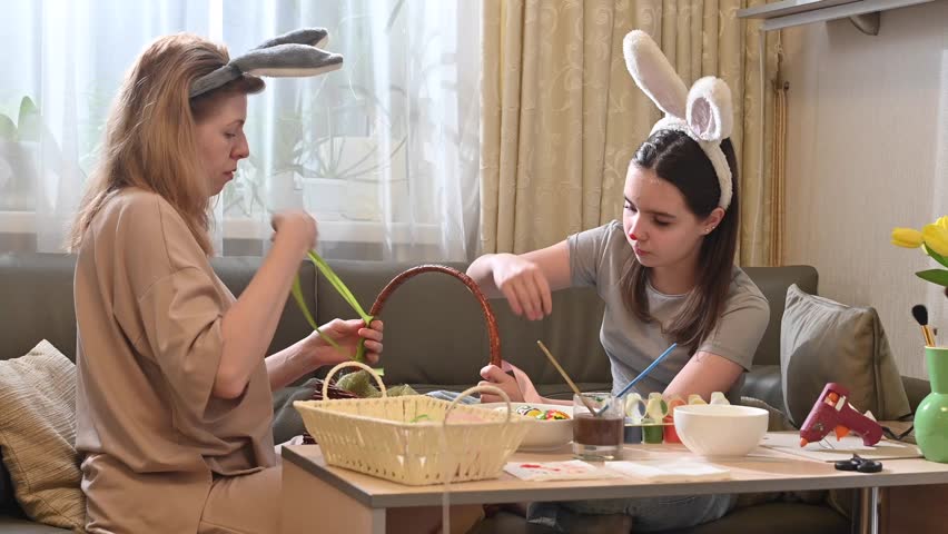 Close-up of two women wrapping a pink ribbon around an Easter basket handle. One holds the basket, the other ties the ribbon. The girl wears bunny ears and has a red nose.