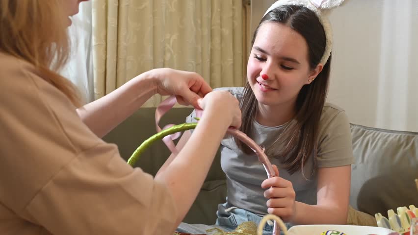 Close-up of two women wrapping a pink ribbon around an Easter basket handle. One holds the basket, the other ties the ribbon. The girl wears bunny ears and has a red nose. Indoor, front view.