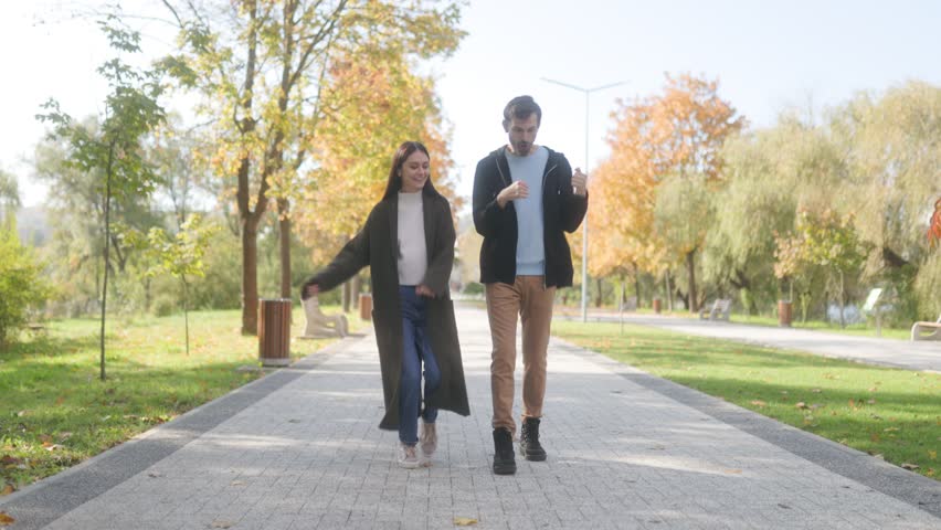 A happy young couple dances playfully on a pathway in an autumn park. Surrounded by fall colors, they enjoy a carefree moment, embracing the joy of being together outdoors.