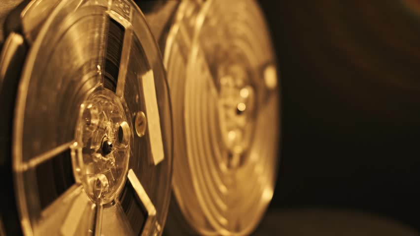 This video shows a close up view of two turning tape reels in an old recorder device in the dim studio space. The tape is on clear bobbins and golden reflections on surface of them.