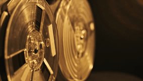 This video shows a close up view of two turning tape reels in an old recorder device in the dim studio space. The tape is on clear bobbins and golden reflections on surface of them. - Powered by Shutterstock - Get 15% off with code: PIKWIZARD15