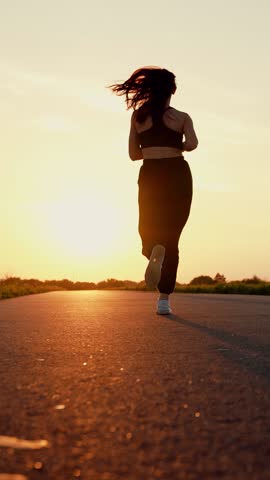 Sporty woman run. Slender girl doing fitness, jogging on road in sun. Jogging outside city, strong young woman runs along asphalt at sunset in summer. Training jogging. Jogger girl breathes outdoor.