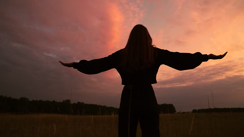 Girl raised her hands to sky in park at sunset. Young woman dreams, plays in park against sky. True faith. Beautiful girl is praying against sky. Religion God, dream. Happy family. Girl looking at sky