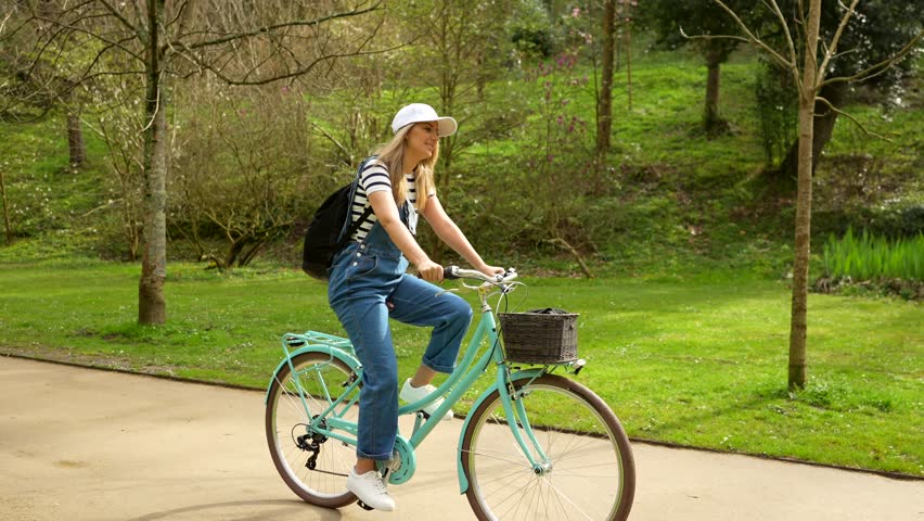 Woman cyclist enjoying leisurely bike ride through verdant park. Wearing casual denim clothing and sun hat. Smiling while traveling along scenic green pathway during sunny spring or summer day