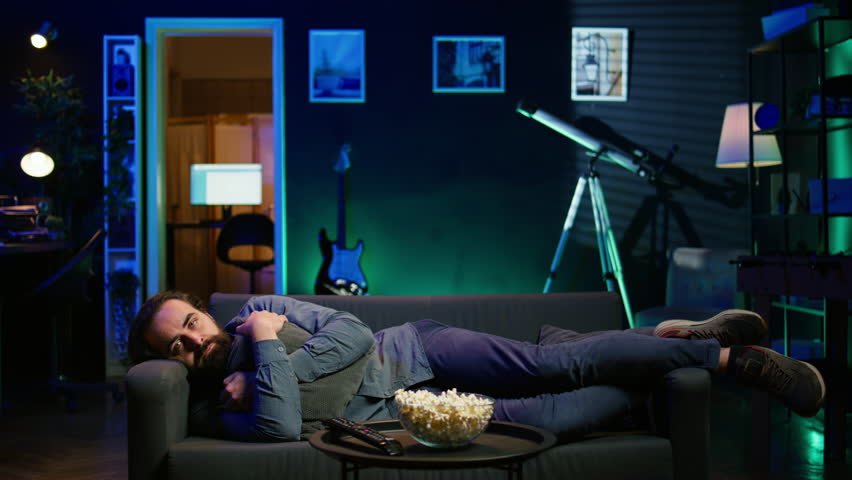 Man laying down hugging pillow watching TV, heartbroken by upsetting movie plot. Sad viewer lounged on couch in RGB illuminated apartment, upset by depressing television show scene, camera B