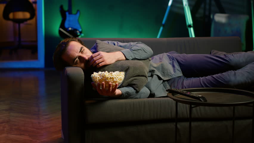 Depressed man at home lounged on couch feeling low, seeking comfort by watching movies on TV and eating popcorn. Glum person laying down, suffering from depression, watching TV to feel better, camera