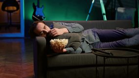 Depressed man at home lounged on couch feeling low, seeking comfort by watching movies on TV and eating popcorn. Glum person laying down, suffering from depression, watching TV to feel better, camera - Powered by Shutterstock - Get 15% off with code: PIKWIZARD15