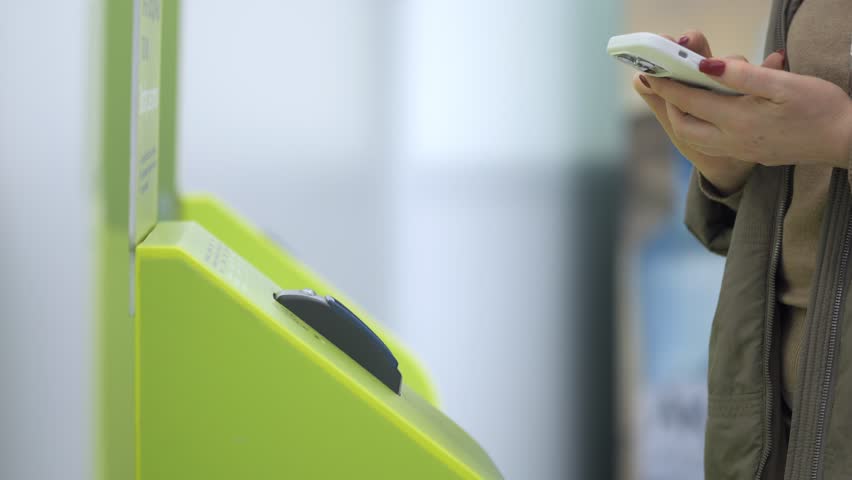 Woman is holding a cell phone in front of a green machine. The machine is a ticket machine, and the woman is likely using it to purchase a ticket