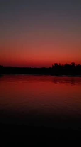 Amazing 20s vertical video of nature, after sunset twilight is reflecting on wavy and rippling river waters in a calm, peaceful, and untroubled state in colorful horizon and dark forest background.