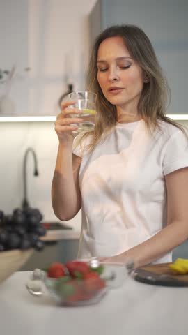 Vertical video, portrait of a woman drinking water from glass standing in the kitchen at home, health care and water balance.