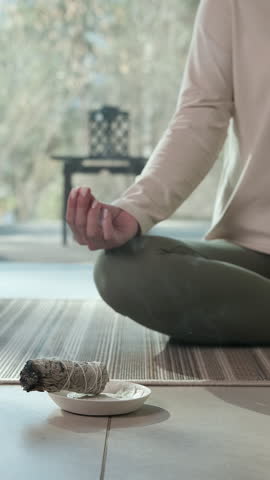 Serene woman practicing yoga and meditation, using white sage smudge stick for spiritual cleansing, creating peaceful atmosphere within comfortable home interior with soft natural light