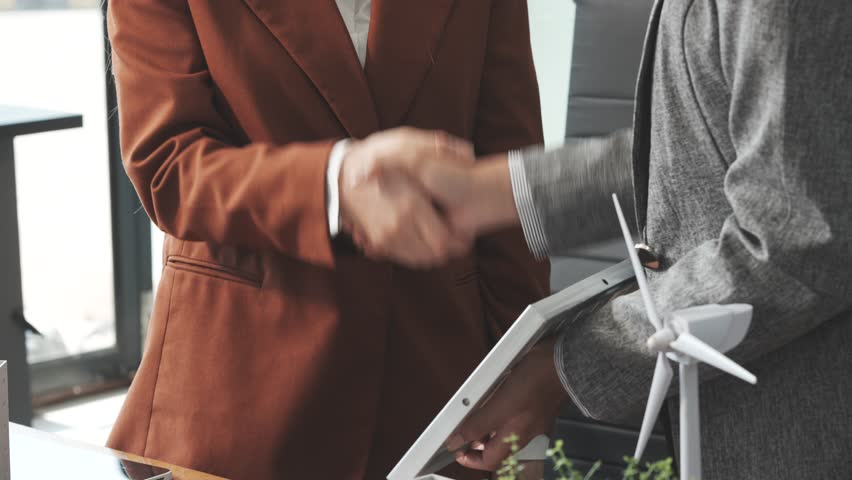 Two businesswomen sit at a desk in a meeting, discussing property development while building a model of city tower. real estate investments, zoning laws, sustainable energy solutions. net zero