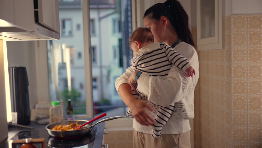 Mother holding baby while cooking in kitchen, stirring food in frying pan, multitasking with child in arms, everyday parenting responsibilities in home setting