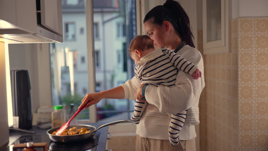 Mother holding baby while cooking in kitchen, stirring food in frying pan, multitasking with child in arms, everyday parenting responsibilities in home setting