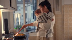 Mother holding baby while cooking in kitchen, stirring food in frying pan, multitasking with child in arms, everyday parenting responsibilities in home setting - Powered by Shutterstock - Get 15% off with code: PIKWIZARD15