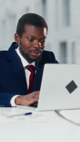 Happy black male worker in modern office opening laptop in workplace, vertical video portrait. Positive businessman monitoring stock rates on display of portable computer, successful business deal