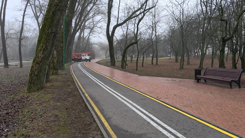 Along the trees on grassy lawns with fallen leaves, a two-lane bike lane with markings and a tile sidewalk are laid out. A tree-trimming autotower truck is on its way. Nearby are lampposts and bench