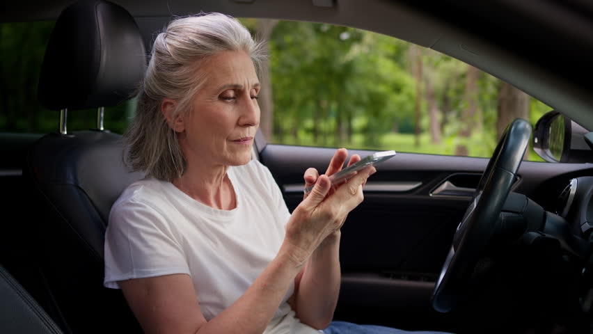 Caucasian grandmother woman female senior driver mature car rental service client user holding mobile phone using artificial intelligence assistant speech app record voice message inside automobile
