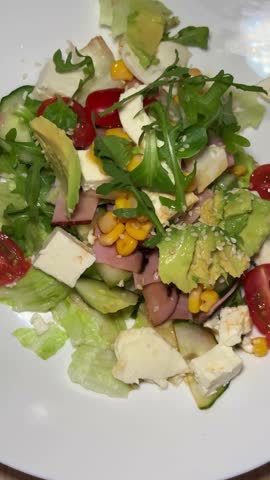 fresh salad with avocado, feta cheese, corn, arugula, cucumber, cherry tomatoes and ham. Light, vibrant and nutritious. Served on a white plate and sprinkled with sesame seeds for a nutty touch.