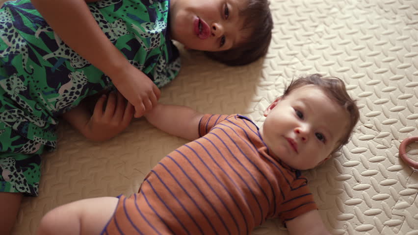 Young boy lying on mat, looking at baby reaching out, gentle sibling interaction, childhood bonding, early family relationship, warm and affectionate moment