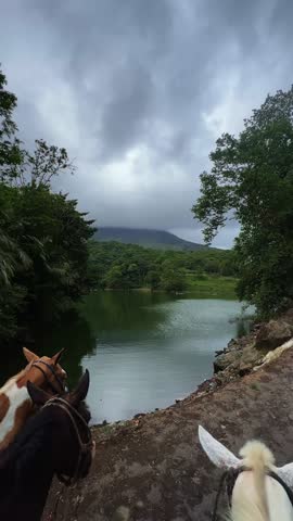 A POV shot from horseback, showing a tropical lake Arenal surrounded by lush rainforest and the Arenal Volcano covered in clouds near La Fortuna, Costa Rica, on a cloudy overcast summer day.